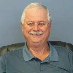 Smiling older man with gray hair wearing a blue shirt, seated against a blue background.