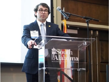 Man speaking at podium during South Alabama event, with microphone and presentation screen in background.